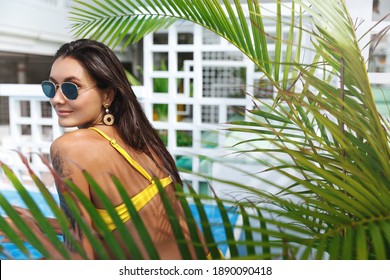 Spa And Tourism. Beautiful Woman Relaxing Near Hotel Swimming Pool In Bikini, Getting Perfect Summer Tan. Female Model Turn Around At Camera, Sunbathing Near Tropical Leaves.