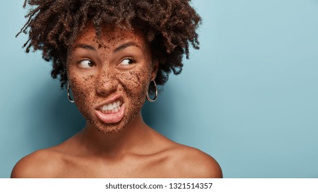 Spa treatments concept. Cropped image of black pretty young woman clenches teeth, frowns face, applies peeling coffee mask, poses topless, focused aside, blank space for your promotional content