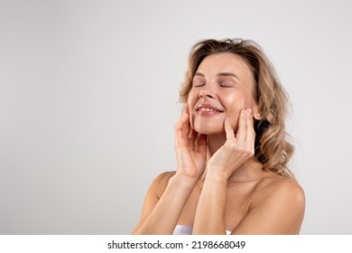 Spa Treatments. Portrait Of Attractive Middle Aged Woman With Beautiful Skin Touching Her Face While Posing Over Light Grey Studio Background, Smiling Lady Wrapped In Bath Towel Enjoying Beauty Care
