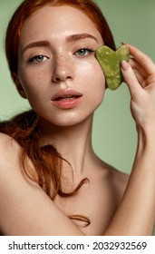 Spa and women beauty. Vertical portrait of redhead girl massage her face with jade massaging scrapper, guasha cosmetology at home, face-lift procedure, standing against green background