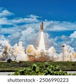 A space shuttle launches into a clear blue sky, leaving a trail of smoke and heat. The landscape below is a mix of green plants and brown soil. . Elements of this image furnished by NASA