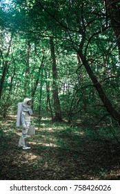 Spaceman wearing uniform is exploring a new place, standing among trees and holding small suitcase. Profile. Copy space on right side