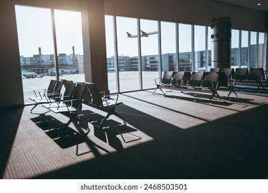 Spacious airport waiting area with empty chairs facing large windows, offering a clear view of the tarmac and a departing airplane. Bright sunlight creates long shadows inside the terminal - Powered by Shutterstock