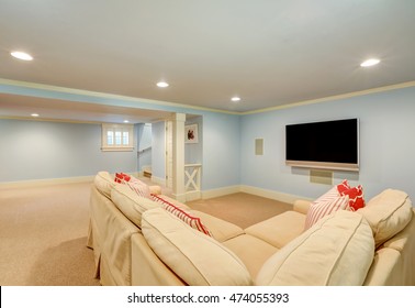 Spacious Basement Living Room Interior In Pastel Blue Tones. Beige Carpet Floor And Large Corner Sofa With TV. Northwest, USA