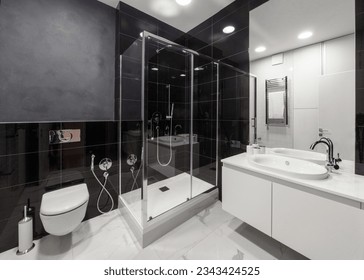 spacious bathroom with black tile on wall and white on floor, shower cabin with glass doors, modern washbowl with faucet and wc toilet with closed lid