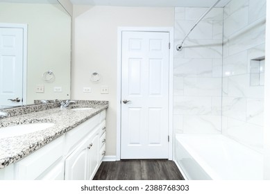  A spacious bathroom with a dual sink vanity, granite countertop, and white cabinetry, leading to a well-lit area with a bathtub and marble tiles - Powered by Shutterstock