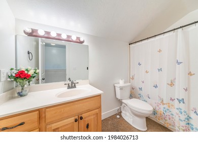 Spacious Bathroom With Vanity Sink And Closed Shower Curtain With Butterflies' Design. There Is A Wooden Vanity And White Countertop With Clear Vase With Red Flowers Near The Electrical Plug.