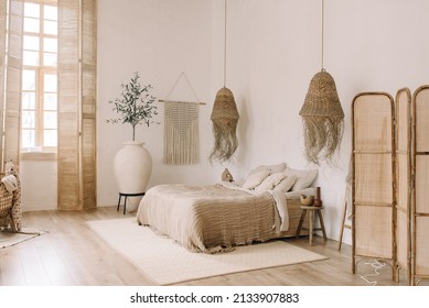 Spacious, bright bedroom with white walls and a large bed in warm boho tones. Straw chandeliers, a large decorative vase, wooden shutters on the windows. - Powered by Shutterstock