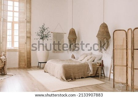 Spacious, bright bedroom with white walls and a large bed in warm boho tones. Straw chandeliers, a large decorative vase, wooden shutters on the windows.