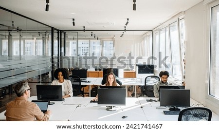 In a spacious and bright office, a diverse team is focused on individual tasks at their workstations, epitomizing the concept of independent productivity within a shared, supportive co-working space