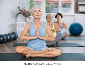 In spacious bright yoga studio, elderly retired woman is engaged in physical education. At class, lady performs half-lotus pose, Ardha Padmasana - Powered by Shutterstock