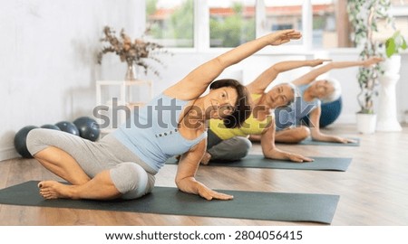 In spacious bright yoga studio, elderly retired woman is engaged in physical education. At class, lady performs half-lotus pose, Ardha Padmasana
