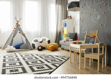 Spacious child room with window, play tent, sack chair, pattern carpet, regale, sofa, small table, chairs and blackboard wall