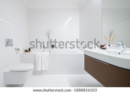 Spacious and elegant bathroom with big bathtub, white tiles, washbasin with wooden cabinet with drawer and big mirror