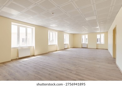 Spacious empty hall or office space featuring beige walls, professional acoustic ceiling tiles, wooden laminate flooring, and several windows with radiators in a new building.