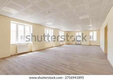 Spacious empty hall or office space featuring beige walls, professional acoustic ceiling tiles, wooden laminate flooring, and several windows with radiators in a new building.