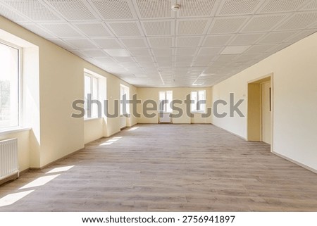 Spacious empty hall or office space featuring beige walls, professional acoustic ceiling tiles, wooden laminate flooring, and several windows with radiators in a new building.