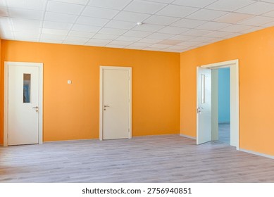 Spacious empty office or classroom interior featuring bright orange walls, wooden laminate flooring, white doors, and a professional acoustic grid ceiling with windows providing natural light