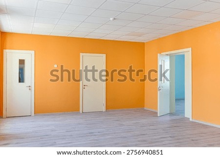 Spacious empty office or classroom interior featuring bright orange walls, wooden laminate flooring, white doors, and a professional acoustic grid ceiling with windows providing natural light