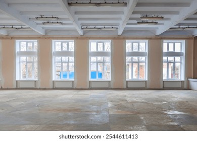 Spacious empty room featuring tall windows, partially finished walls, and concrete floor. Natural light floods through the windows, giving open, airy feel. Ideal for renovation or remodeling projects
