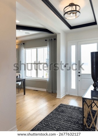 Spacious entryway featuring a coffered ceiling, patterned black rug, and wide windows with curtains. Light walls and natural wood flooring give the space an inviting look.