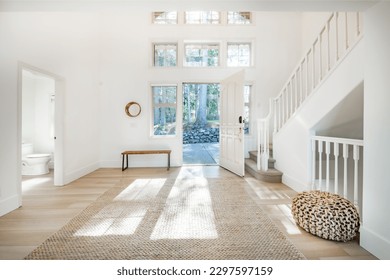 spacious foyer entry to an entry with dappled sunshine jute ottoman wooden bench open door with bright light shining in to a dual white wood railed stairway - Shutterstock ID 2297597159