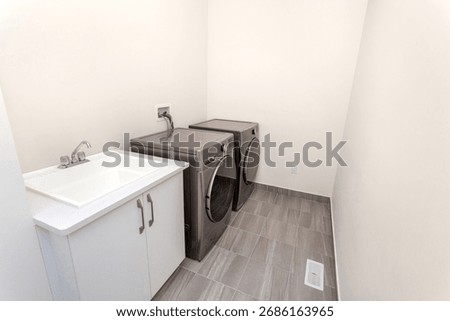 Spacious laundry room with front-load washer and dryer, utility sink cabinet, gray tile floor, and neutral walls—clean and ready to use.