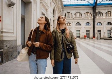 In a spacious, light, high-ceilinged train station, two White casually dressed women in their 20s, a blonde and a brunette, are standing close to each other and looking up, at the architecture.
