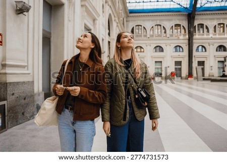 In a spacious, light, high-ceilinged train station, two White casually dressed women in their 20s, a blonde and a brunette, are standing close to each other and looking up, at the architecture.