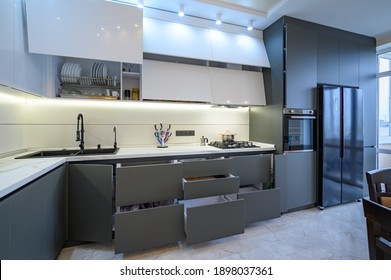 Spacious Luxury White And Dark Grey Modern Kitchen Interior With Dining Table, Most Furniture Drawers And Doors Are Open