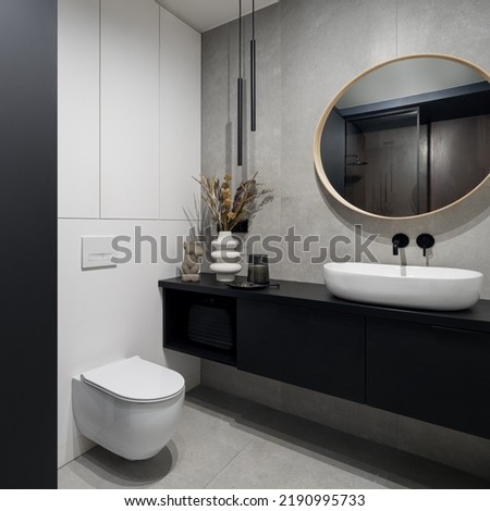 Spacious and modern bathroom with concrete gray floor and wall tiles, black furniture, big round mirror, oval washbasin, toilet and stylish decorations