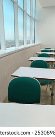 Spacious modern classroom with rows of empty desks and green chairs, featuring large windows offering a panoramic city view