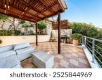 Spacious outdoor terrace with wooden pergola, wicker sofa, coffee table, and grill. Tiled floor, potted plants, latticework, and surrounding greenery create relaxing atmosphere. outdoor living