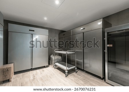 A spacious professional kitchen area features stainless steel refrigerators arranged neatly against the walls. A service cart stands ready for use, ensuring efficient food storage and preparation
