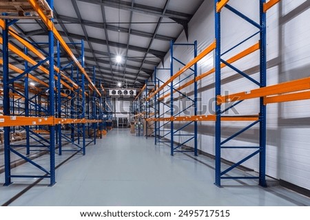 Spacious Warehouse Interior Featuring Empty Blue and Orange Shelving Units Under Bright Lighting.
A large, empty warehouse showcases rows of shelving units. The high ceilings and bright overhead light
