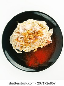 Spaghetti Carbonara With Bacon, Creamy Sauce, Fried Onion Sprinkle With Red Paprika Powder Sprinkle On A Black Ceramic Plate On A White Background