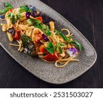 Spaghetti with chicken, tomatoes, herbs and edible flowers is served on a gray plate lying on a dark wooden table