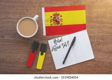 Spain national flag, notebook with the inscription hello, how are you in spanish, two markers, cup of coffee on brown wooden desktop, study concept
