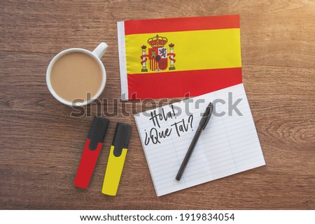 Spain national flag, notebook with the inscription hello, how are you in spanish, two markers, cup of coffee on brown wooden desktop, study concept