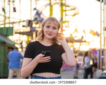 A Spanish Blonde Woman Holding A Phone Near A Rollercoaster In The Park During The Sunset