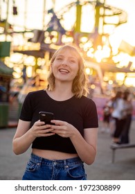 A Spanish Blonde Woman Holding A Phone Near A Rollercoaster In The Park During The Sunset