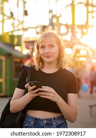 A Spanish Blonde Woman Holding A Phone Near A Rollercoaster In The Park During The Sunset