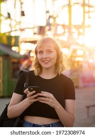 A Spanish Blonde Woman Holding A Phone Near A Rollercoaster In The Park During The Sunset