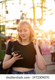 A Spanish Blonde Woman Holding A Phone Near A Rollercoaster In The Park During The Sunset