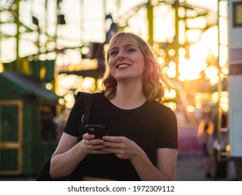 A Spanish Blonde Woman Holding A Phone Near A Rollercoaster In The Park During The Sunset