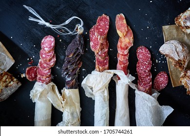 Spanish Dry Sausages In Craft Paper With Bread On Black Wooden Background, Assortment Of Delicious Deli Meats, Salami And Pepperoni  