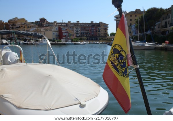 the Spanish flag on the flagpole of\
the yacht against the background of the port and the picturesque\
colorful Spanish houses and the blue sea in sunny\
weather