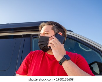 A Spanish Man With A Black Fabric Mask Talking On His Phone With A Car In The Background