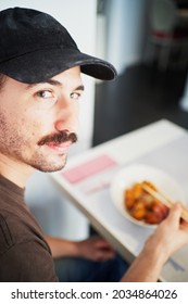 Spanish Man Wearing Cap Looks At Camera While Eating Asian Food With Chopsticks