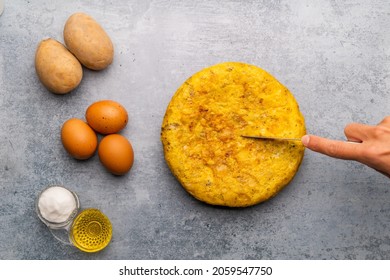 Spanish Omelette With Ingredients On The Side On Concrete Texture Background  Hand Cutting A Portion Of The Tortilla  Overhead View 